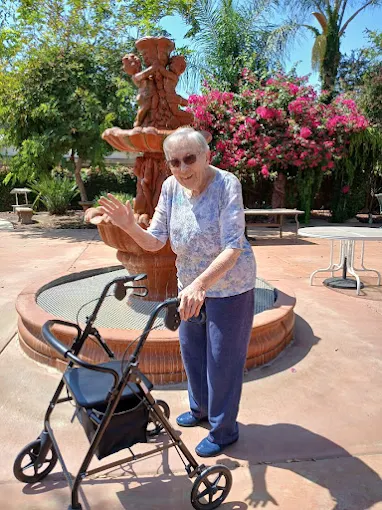 Happy senior woman with walker waving by a decorative fountain in the beautiful garden area of the Glory Days Assisted Living facility.