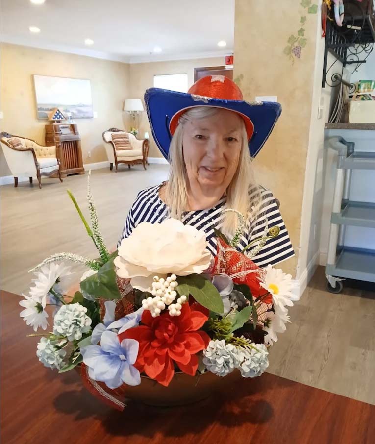 Happy memory care resident wearing a festive patriotic hat and holding a flower arrangement during holiday celebrations at a senior living home