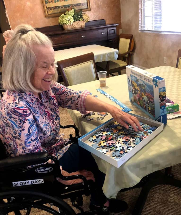 Smiling elderly woman in a wheelchair engaging in cognitive stimulation by solving a jigsaw puzzle at Glory Days memory care specialists.