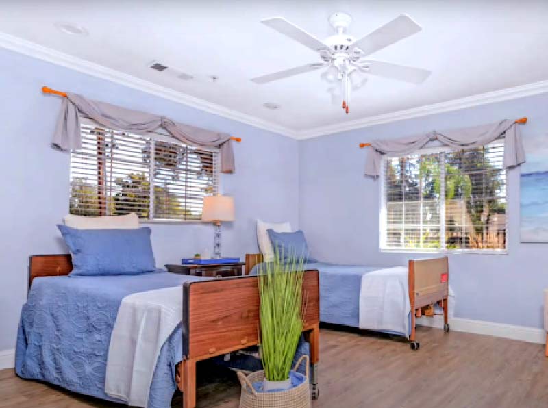 A bright, welcoming shared resident bedroom with two comfortable beds, cozy blue bedding, a ceiling fan, and large windows providing natural light at our senior care facility.